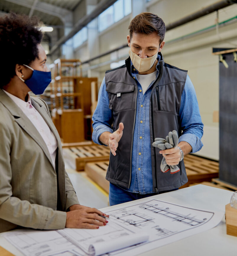 Woodworker and black businesswoman wearing protective face masks while examining blueprints in a factory.