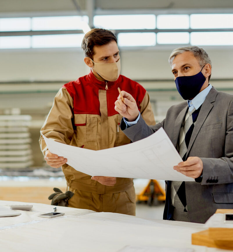 Mid adult businessman and manual worker analyzing plans while working at carpentry workshop during coronavirus pandemic.